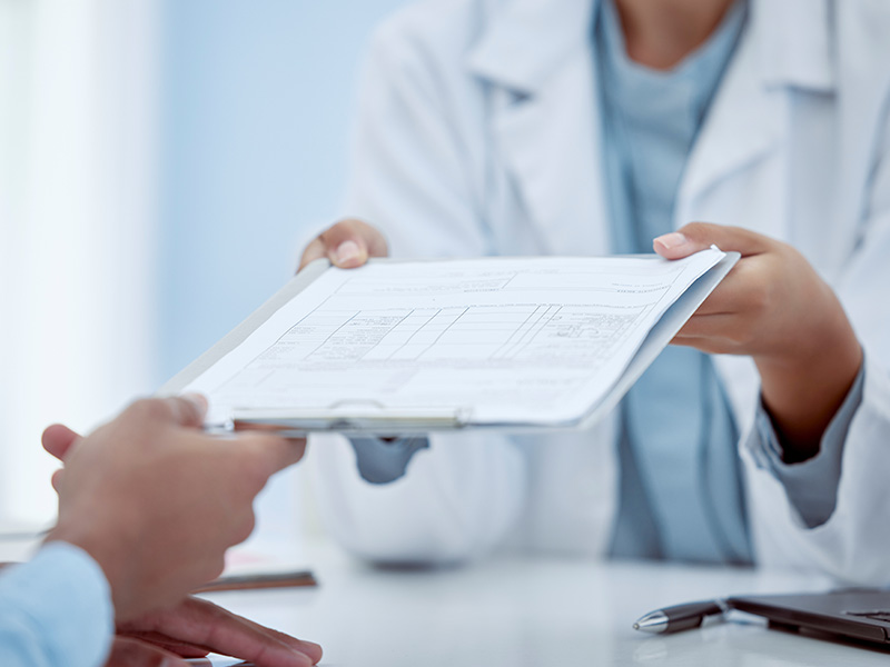 The image features two individuals at a table, with one person holding an open document and another handing over a clipboard, set against a backdrop of a medical office environment.