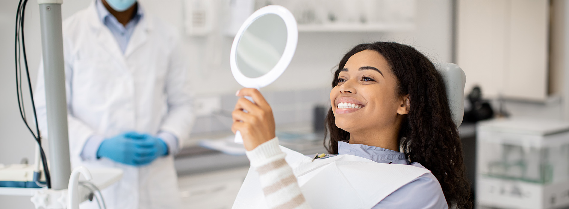 A person is seated in a dental chair, receiving care from a dental professional who stands behind them.